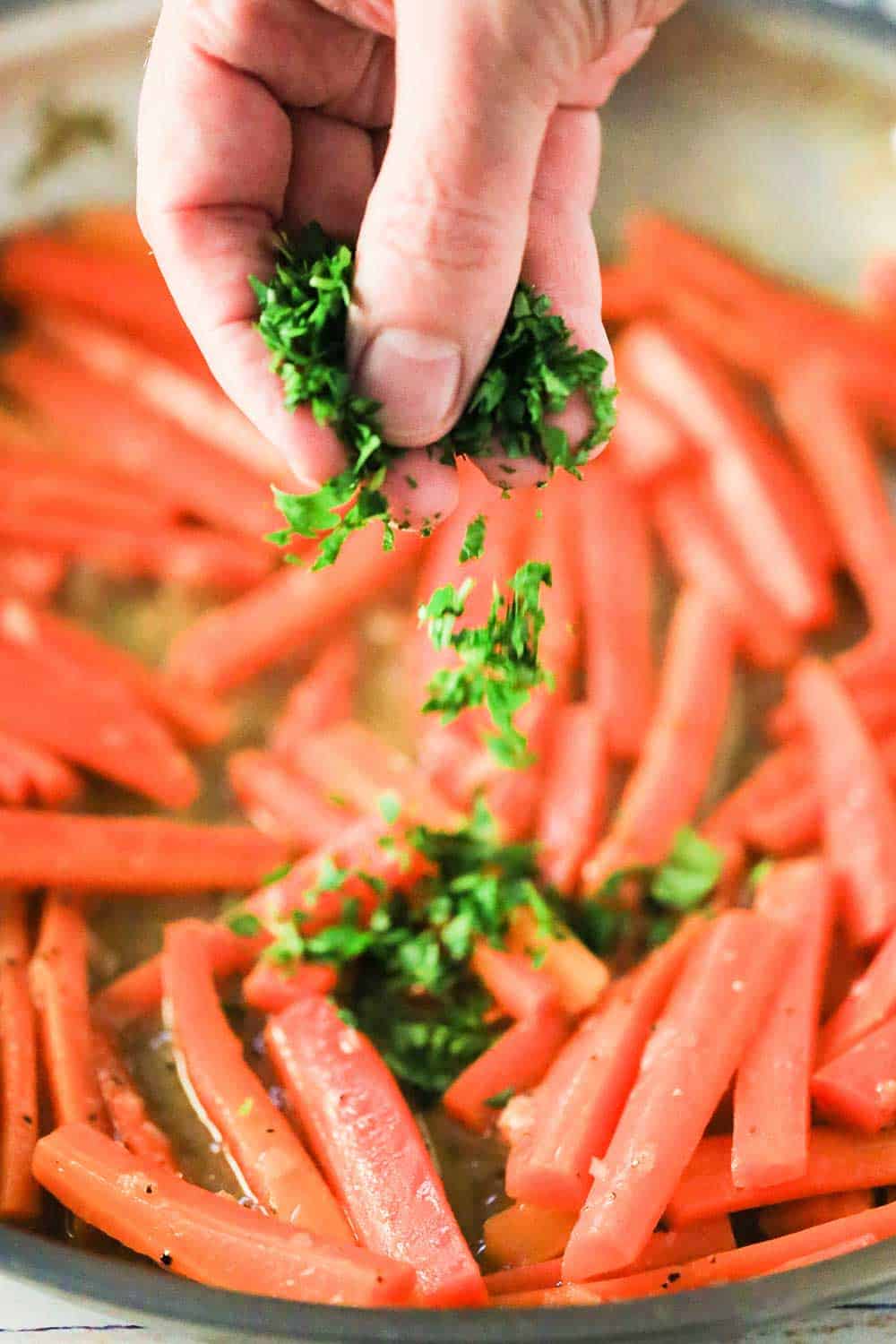 A hand dropping fresh chopped parsley into a skillet of maple braised carrots. 