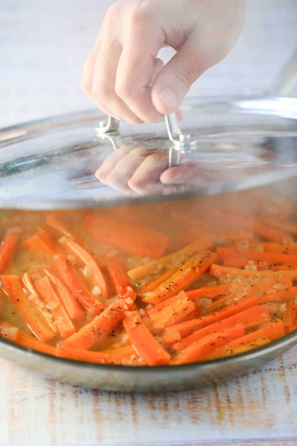 A hand lifting a lid from a large silver skillet filled with braised carrots and steam is escaping. 