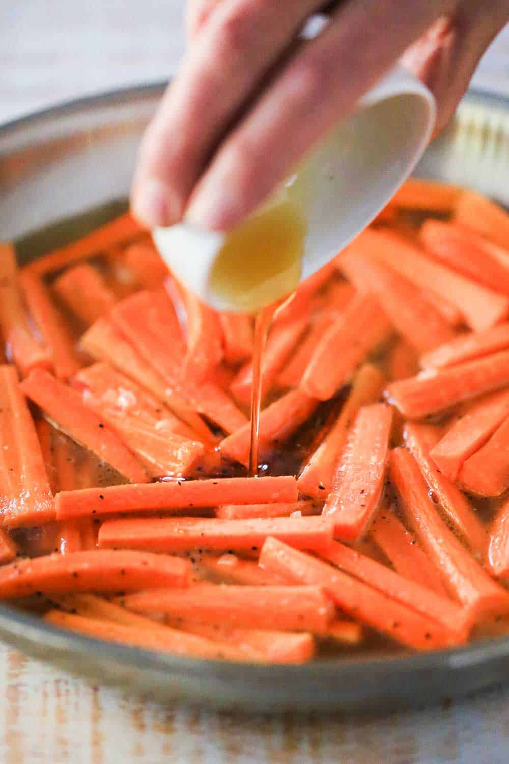 A hand pouring maple syrup from a small white bowl into a skillet with sautéed carrots in it. 