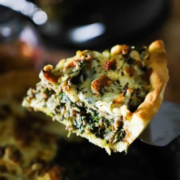 A slice of deep-dish pizza with sausage and spinach being lifted from the pan with a spatula.