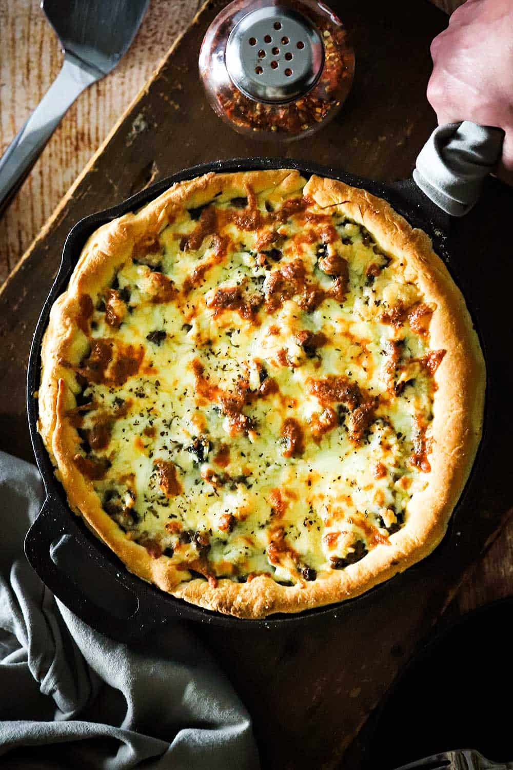 A hand holding the handle of a cast-iron skillet that is filled with deep-dish sausage and spinach pizza that has just been placed on a wooden table. 