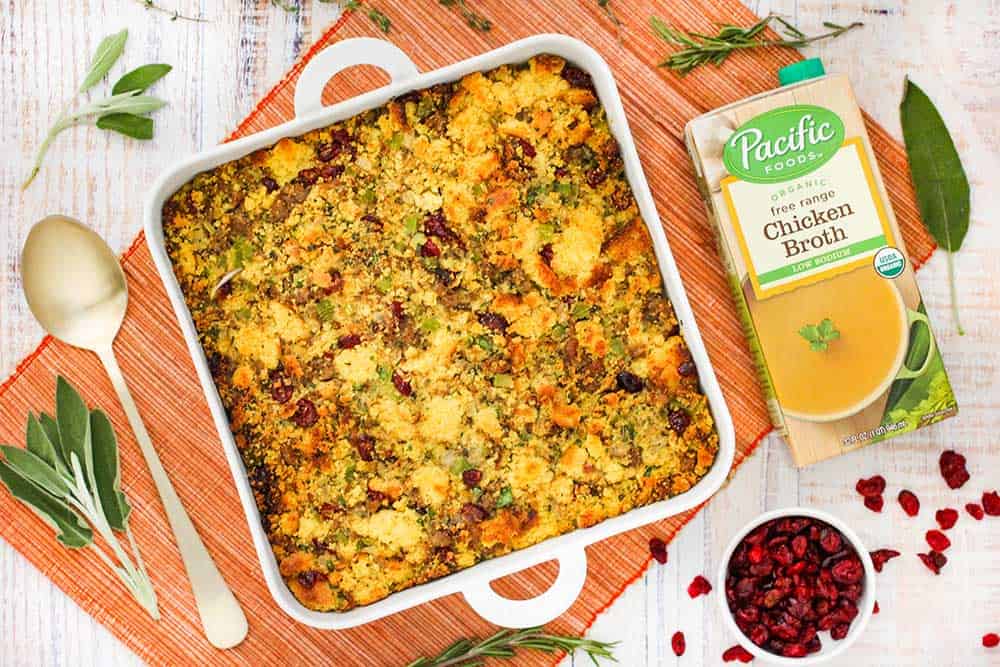 An overhead view of a square dish of cornbread dressing with sausage and cranberries next to a box of chicken broth and a large spoon and herbs. 