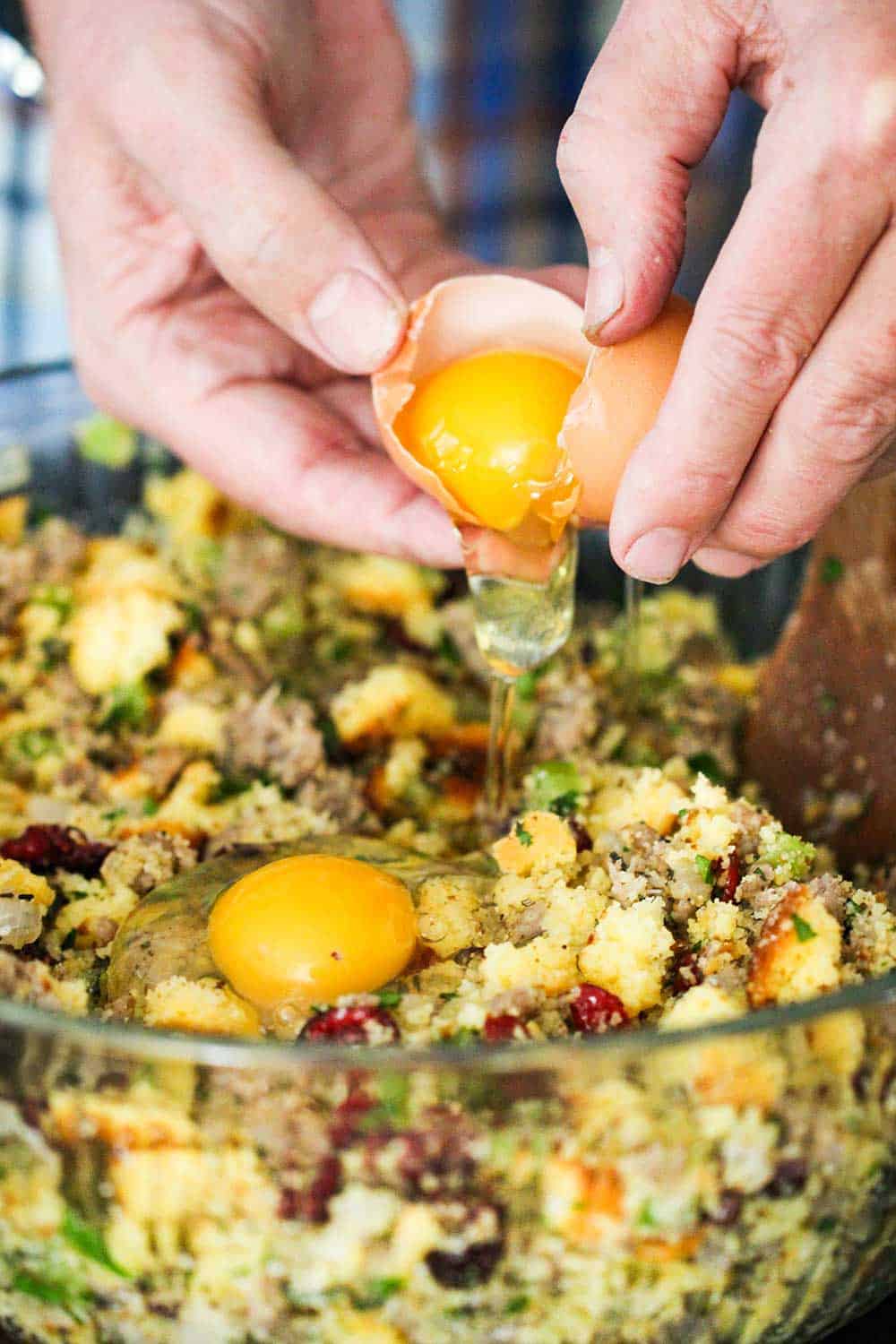 Two hands cracking an egg into a large glass bowl of cornbread dressing with sausage and cranberries. 