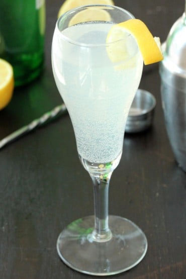 A Champagne flute filled with a French 75 cocktail with a lemon twist on it next to a cocktail shaker.