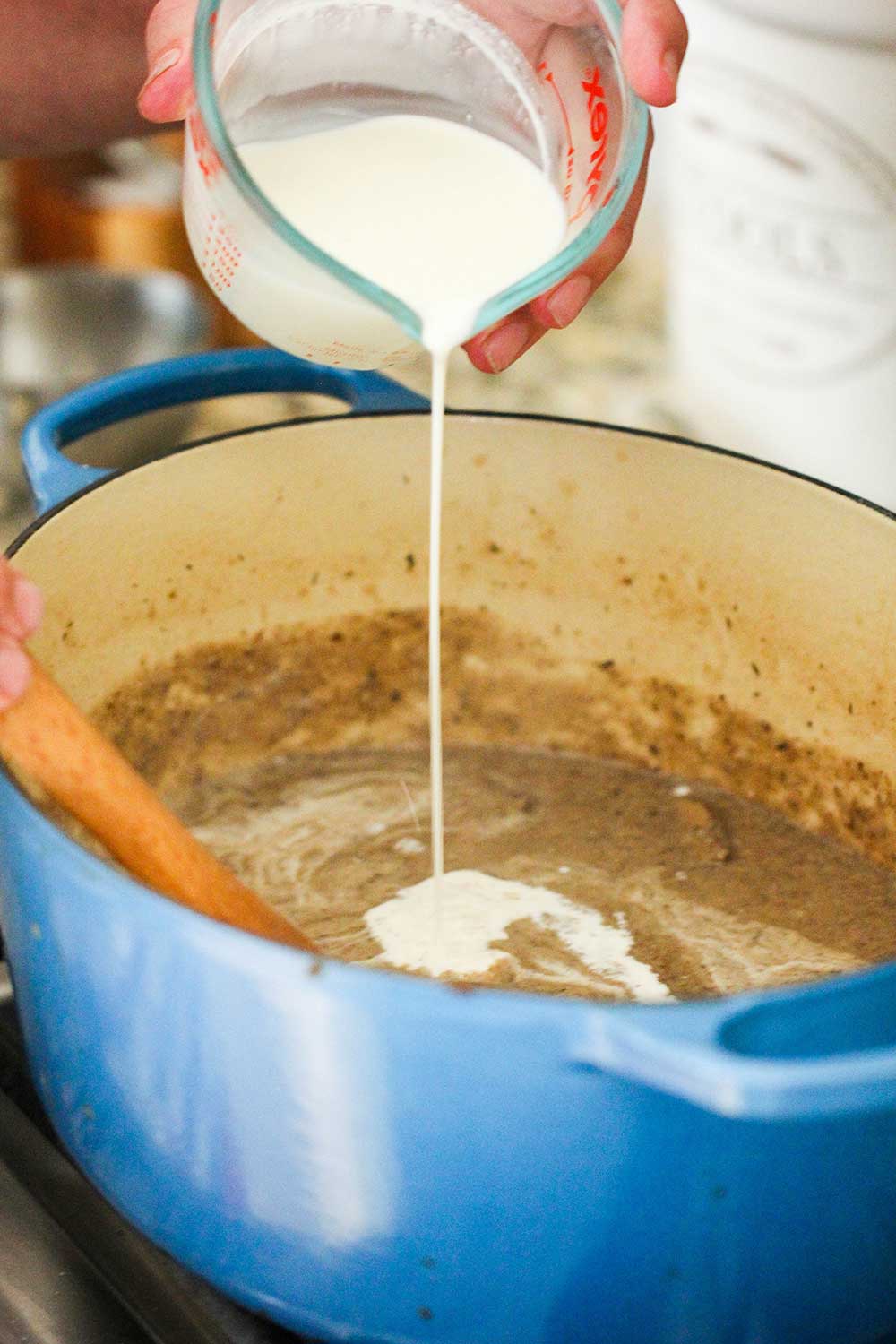 A cup of cream being poured into a large blue Dutch oven of sautéed mushroom and sauce. 