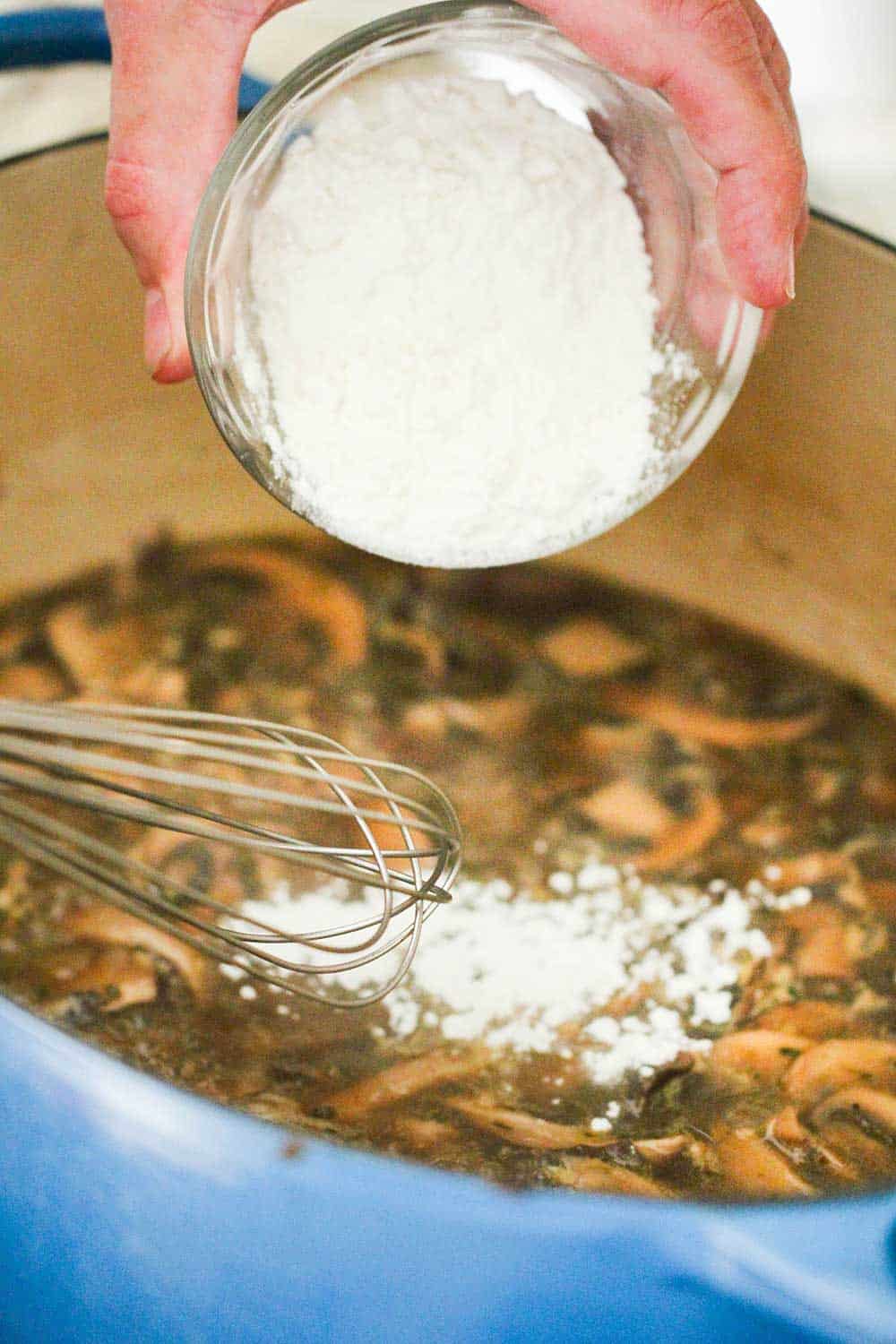 A hand dumping flour into a Dutch oven with sautéed mushrooms for cream of mushroom soup. 