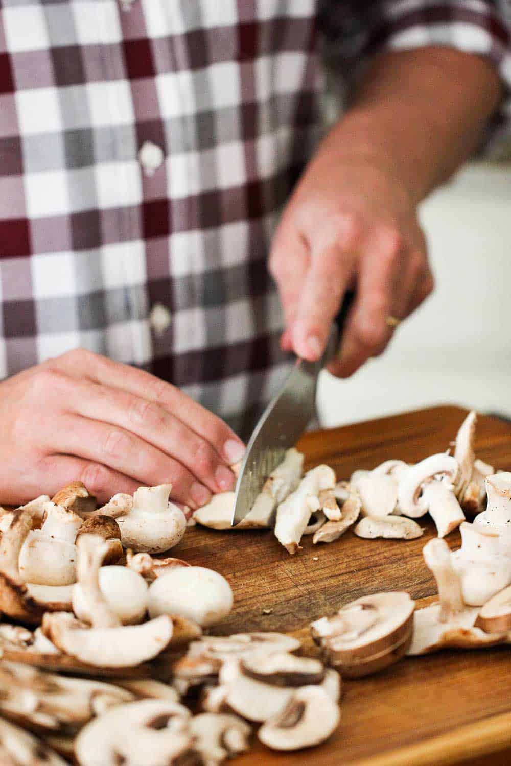 Fresh mushrooms being sliced on a wooden cutting board with two hands and chef's knife. 