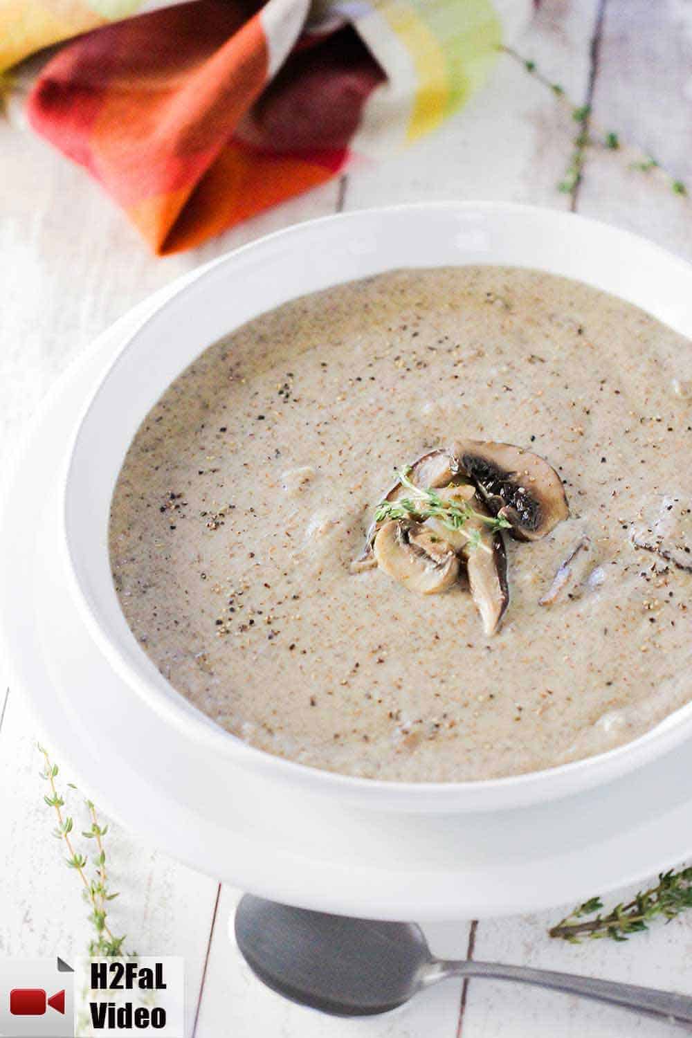 A white bowl filled with cream of mushroom soup on a white background with a spoon and a sprig of thyme next to it. 