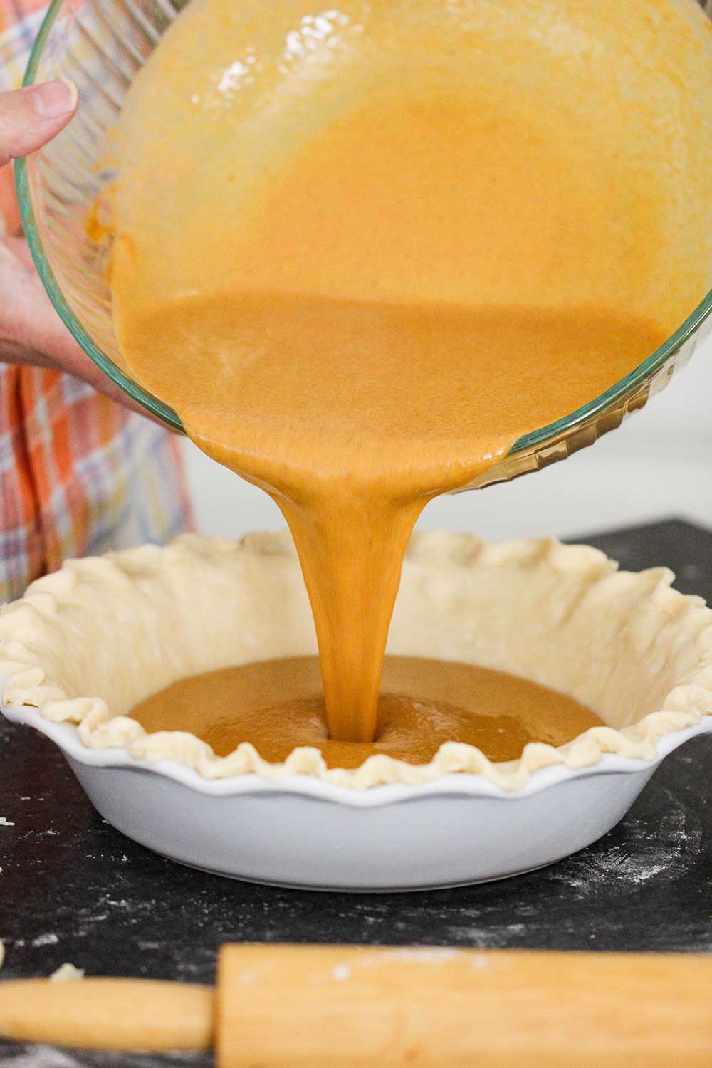 Two hands pouring pumpkin pie batter into a pie dish lined with pie dough. 