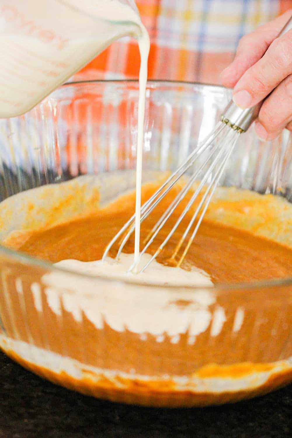 A hand pouring in cream into pumpkin pie batter in a large glass bowl. 