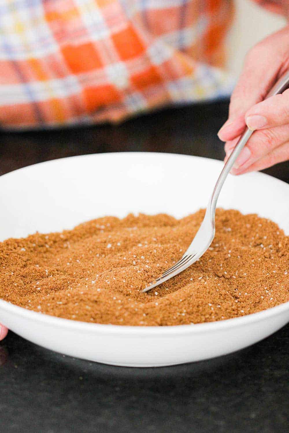 A hand using a fork in a white bowl to blend together spices for classic pumpkin pie. 