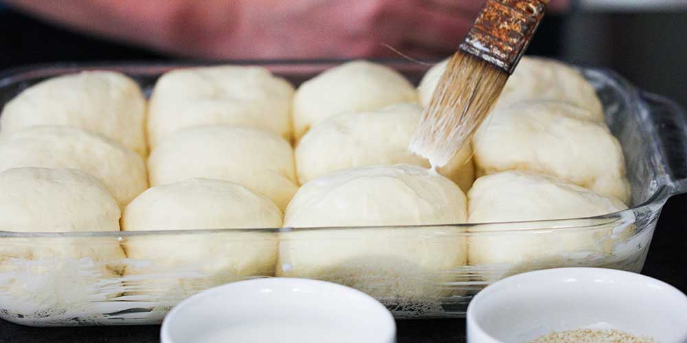 A hand using a small brush to add milk over the tops of doughs of rolls in a glass baking dish. 