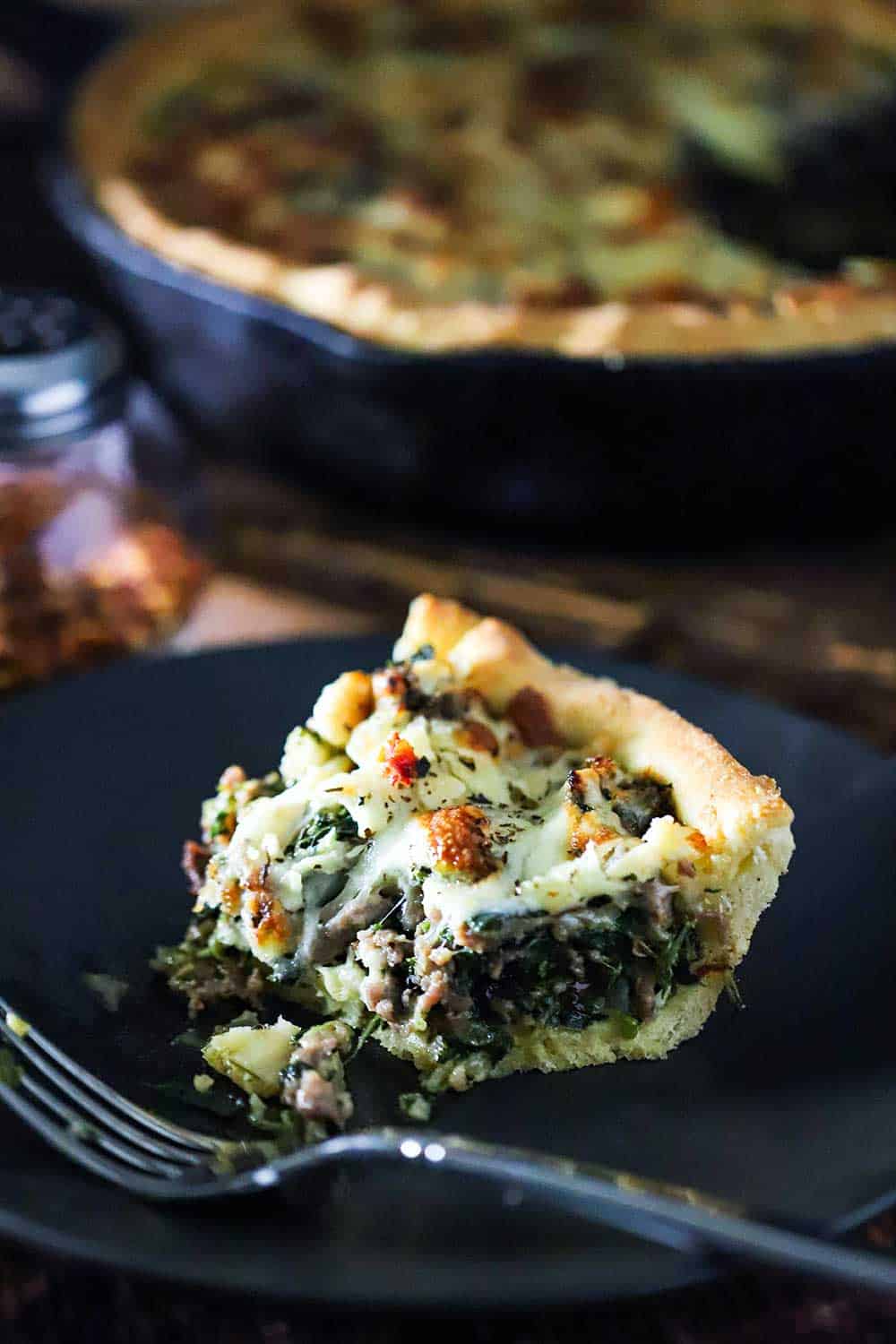 A slice of deep-dish pizza with sausage and spinach with a bite taken out of it sitting on a blue plate with a fork next to it. 