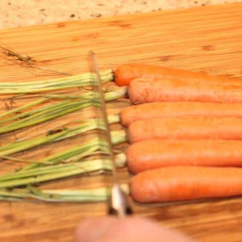 A hand holding a knife trimming the carrot tops on a wood cutting board