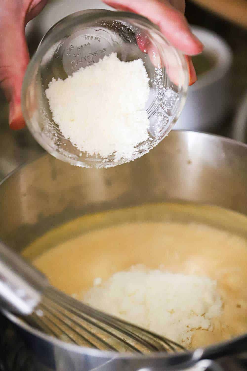A hand dumping 1/4 cup of grated Parmesan cheese into a saucepan filled with a bechamel sauce. 