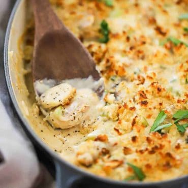 A wooden spoon sitting in casserole dish filled with cooked oyster casserole, with one serving missing from the dish.