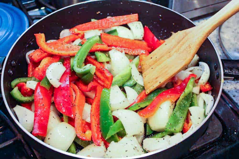 Red and green peppers and sliced onions simmering in a large black skillet with a wooden spoon. 