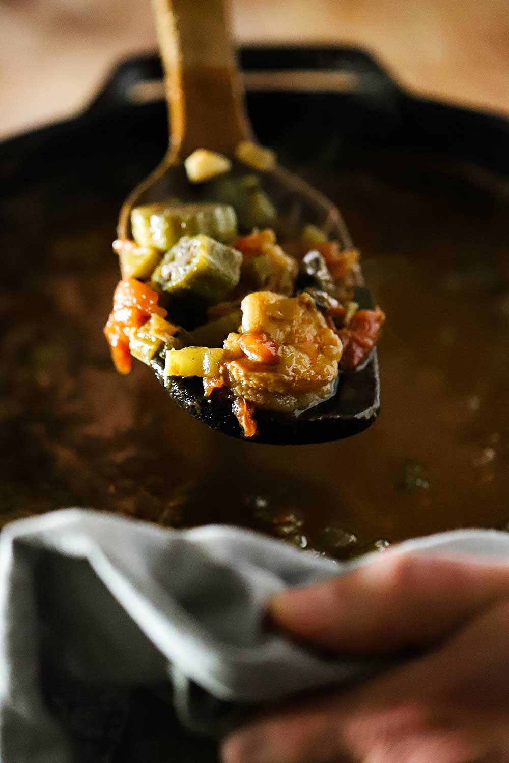 A large wooden spoon being lifted up out of a pot of shrimp and okra gumbo holding a helping of the dish. 
