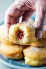 A hand holding a jelly and custard doughnut with a bite taken out of it.