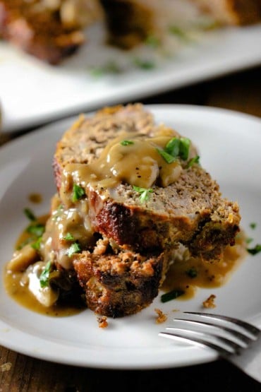 Two slices of best-ever meatloaf sitting on top of each other on a white plate with mushroom gravy on top.