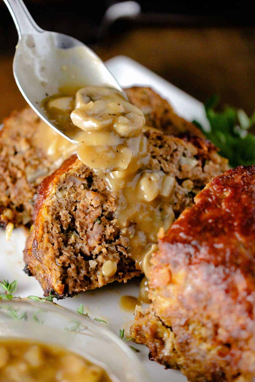 A spoon drizzling mushroom gravy over slices of a meatloaf on a white platter. 