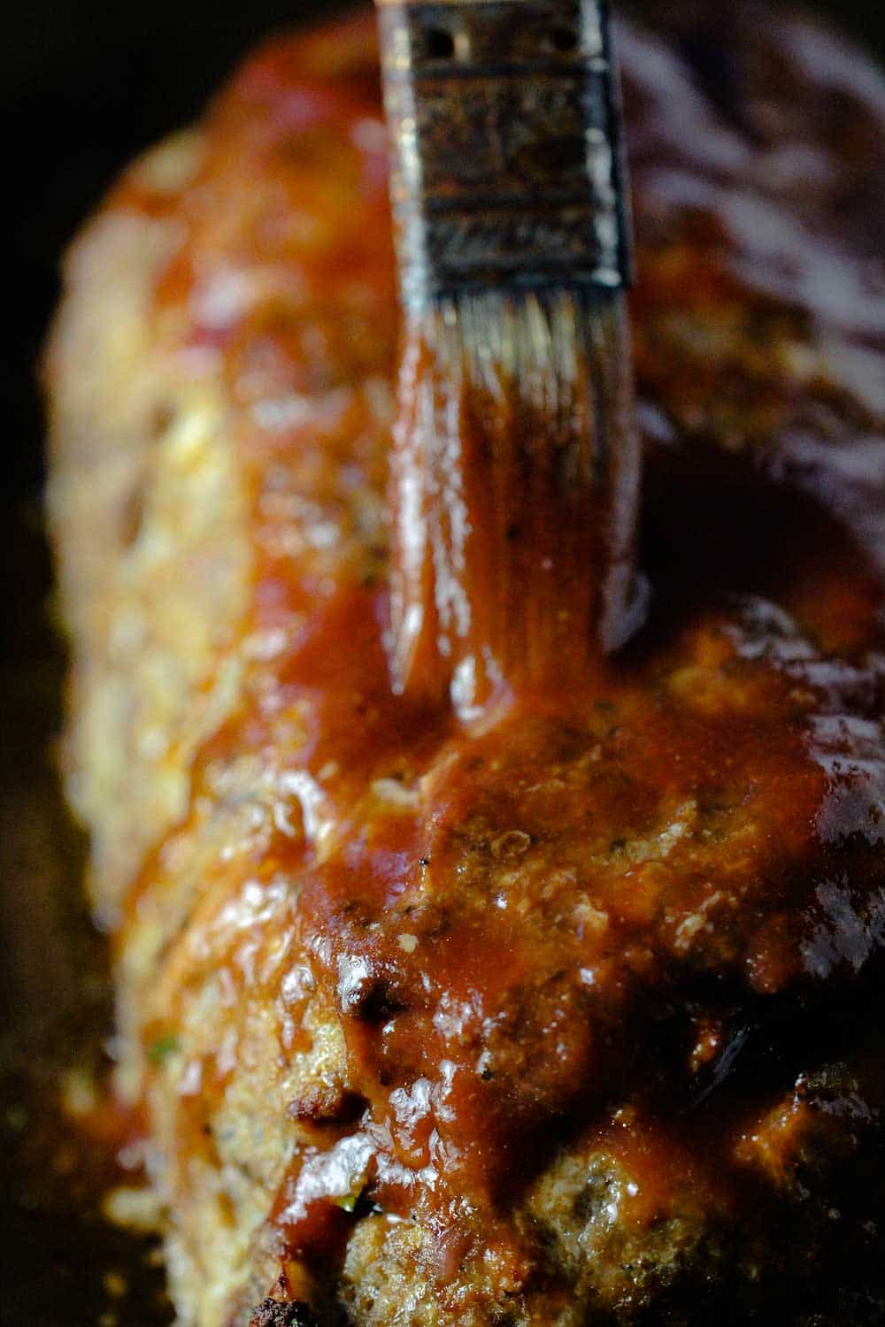 Une main utilise un pinceau pour appliquer une sauce tomate sur le dessus du meilleur pain de viande de tous les temps, partiellement cuit. 