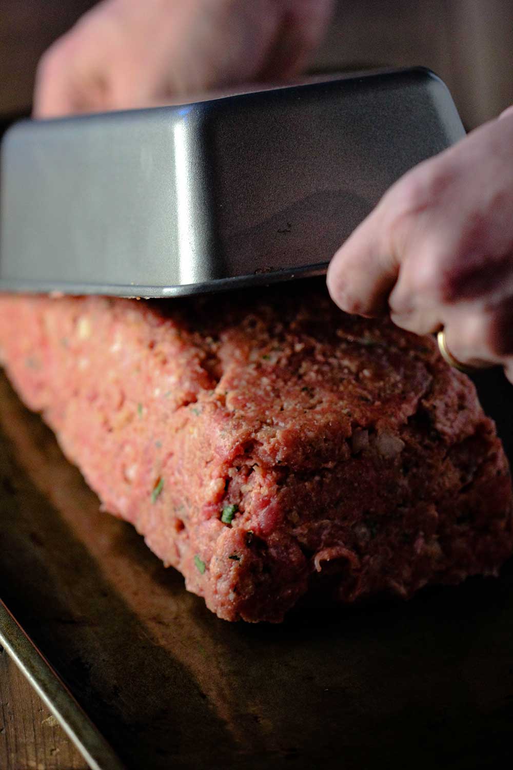 Two hands lifting a metal loaf pan away from an uncooked meatloaf on a baking sheet. 