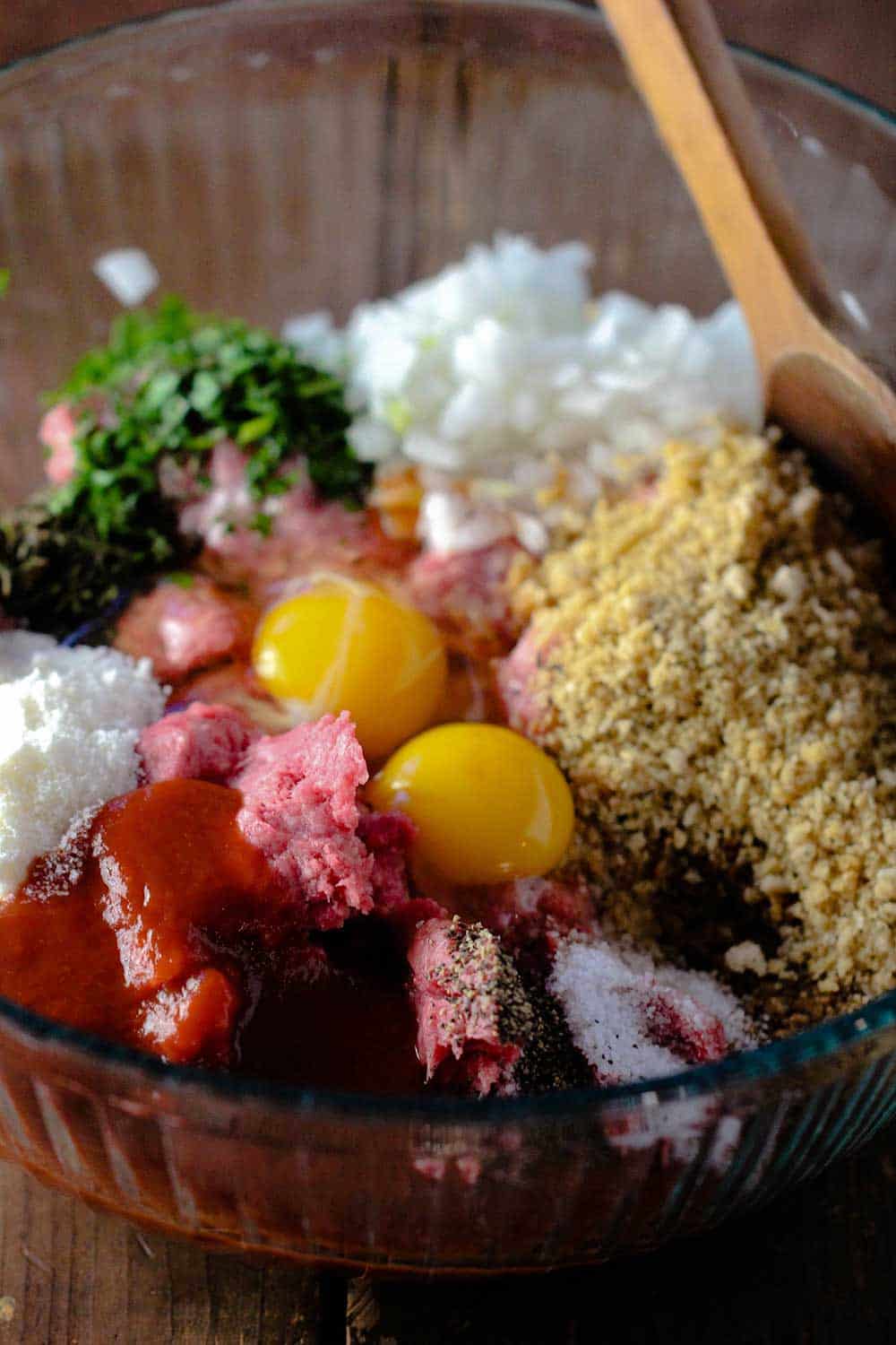 A glass bowl holding ground meat with eggs, bread crumbs, cheese and fresh herbs. 