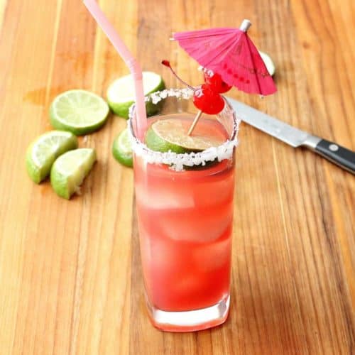 A tall glass holding a Mexican Sea Breeze Cocktail with a pink straw on a wood cutting board with lime slices