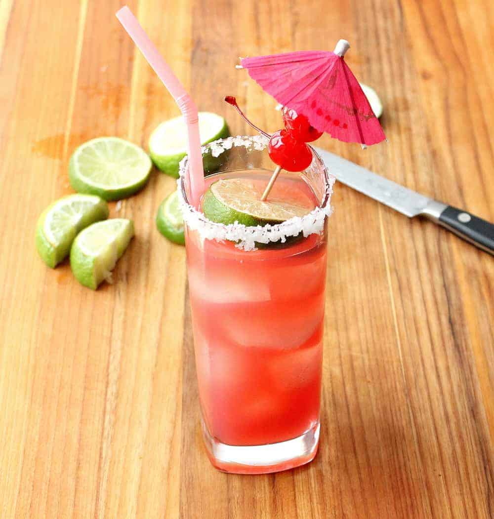 A tall glass holding a Mexican Sea Breeze Cocktail with a pink straw on a wood cutting board with lime slices