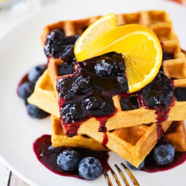 A stack of buttermilk waffles topped with a blueberry sauce and a slice or orange all on a white plate with a fork next to it.