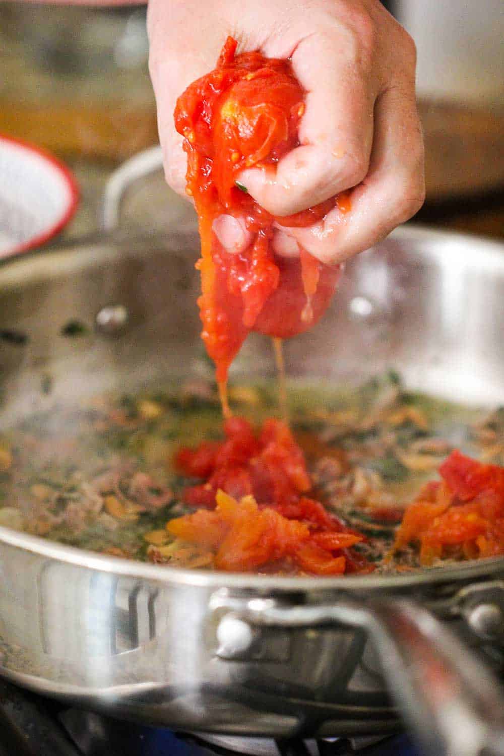 A hand squeezing whole tomatoes into a large skillet for pasta pomodoro. 