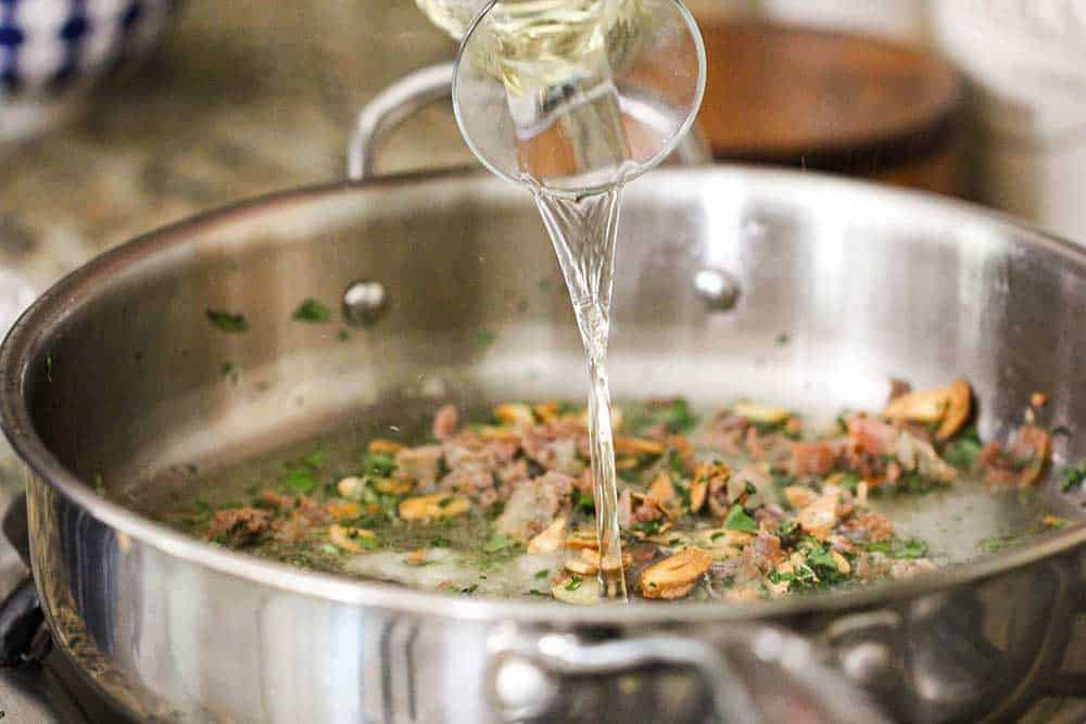 White wine being poured into a large saucepan filled with sautéed garlic, prosciutto and herbs. 