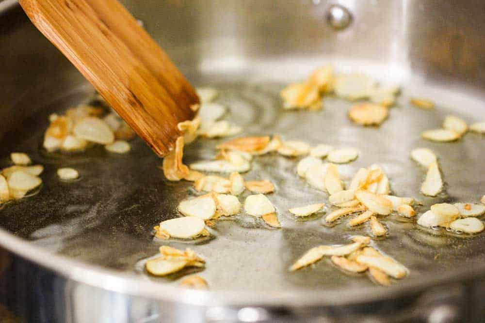 Garlic slivers cooking in a sliver pan with olive oil and a wooden spoon. 