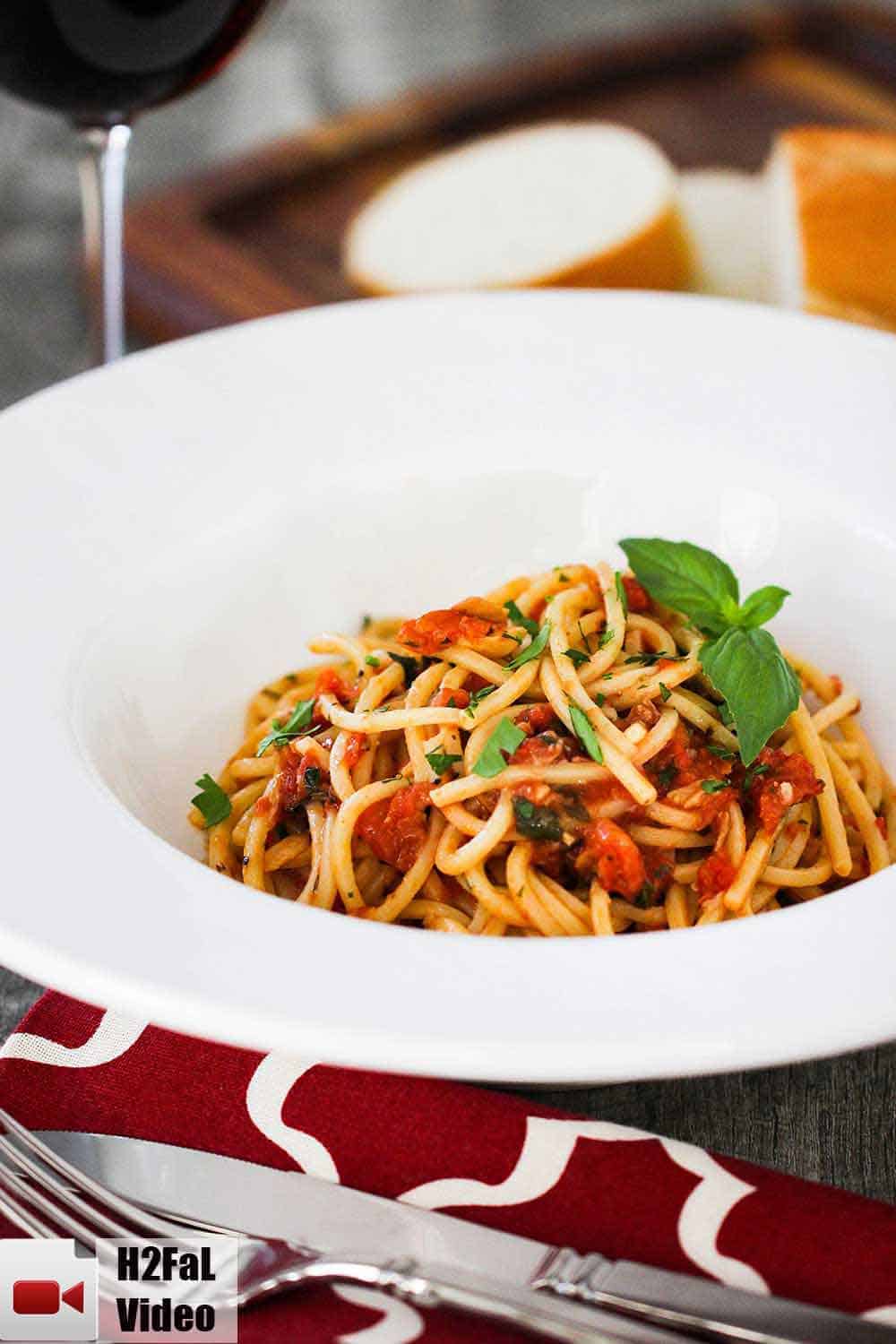 A large white bowl filled with fresh pasta pomodoro next to a glass of red wine and a red-checkered napkin. 