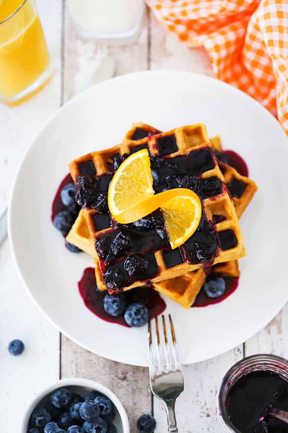 A stack of buttermilk waffles topped with a blueberry sauce and a slice or orange all on a white plate with a fork next to it.