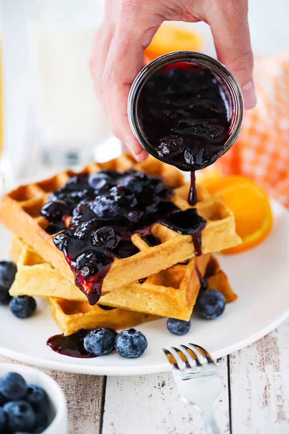 A hand pouring a blueberry sauce over a stack of buttermilk waffles on a white plate next to a fork. 