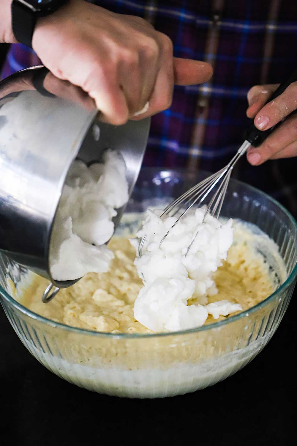 A person transferring beaten egg whites into a glass bowl filled with buttermilk waffles batter. 