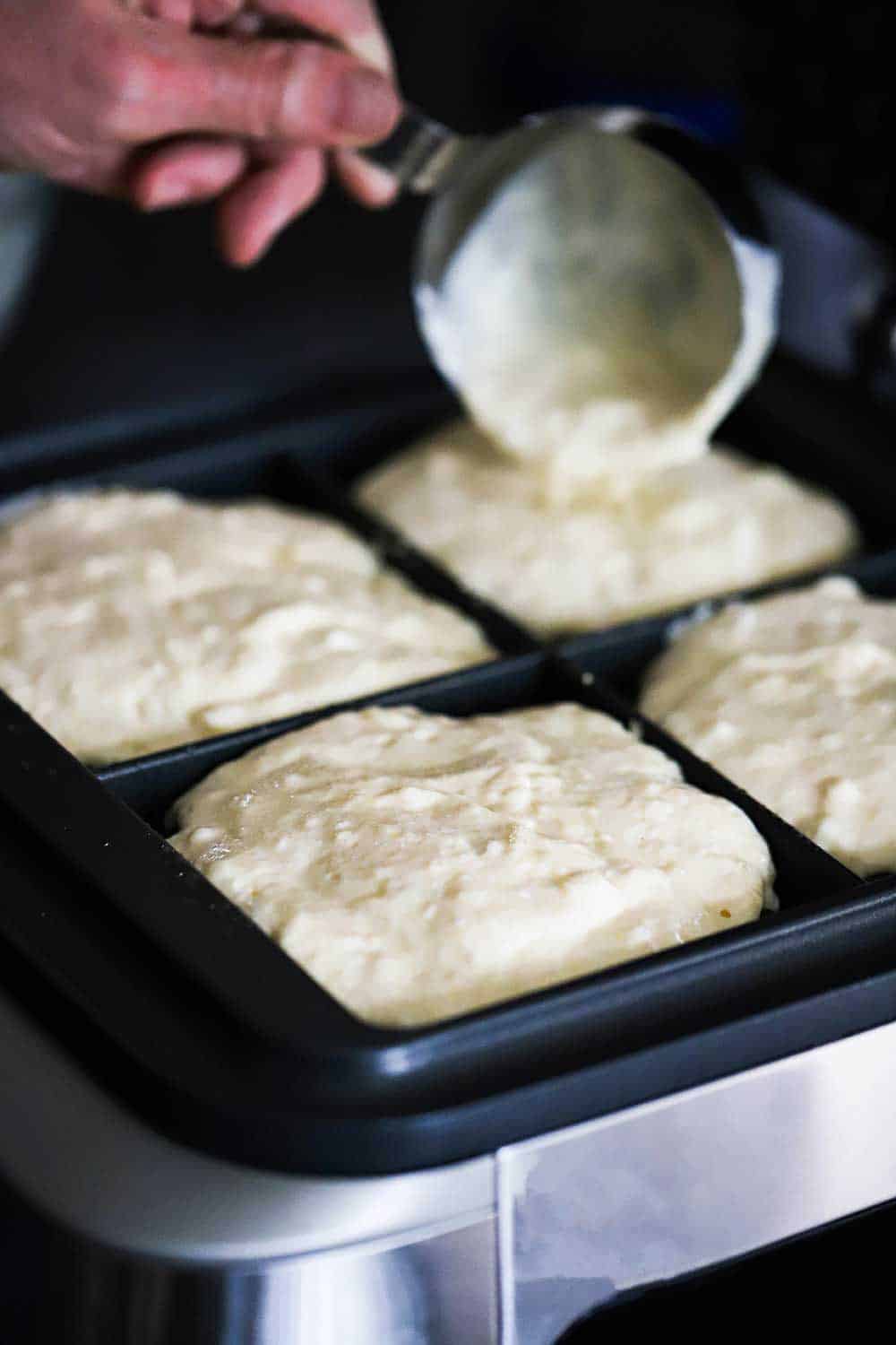 A person using a metal measuring cup to transfer waffle batter into a square waffle maker. 