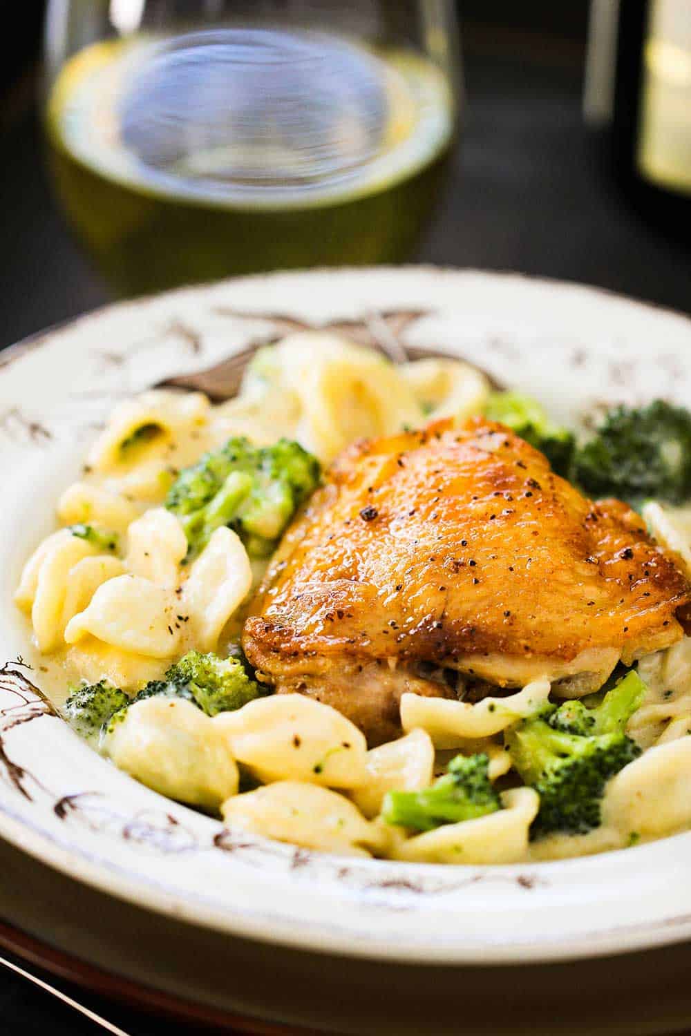 An antique bowl with chicken and broccoli alfredo in it, next to a glass of white wine. 