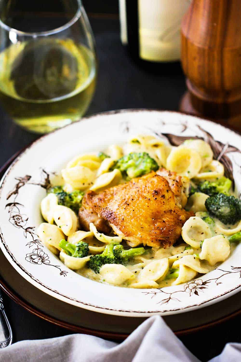 An antique bowl with chicken and broccoli alfredo in it, next to a glass of white wine. 