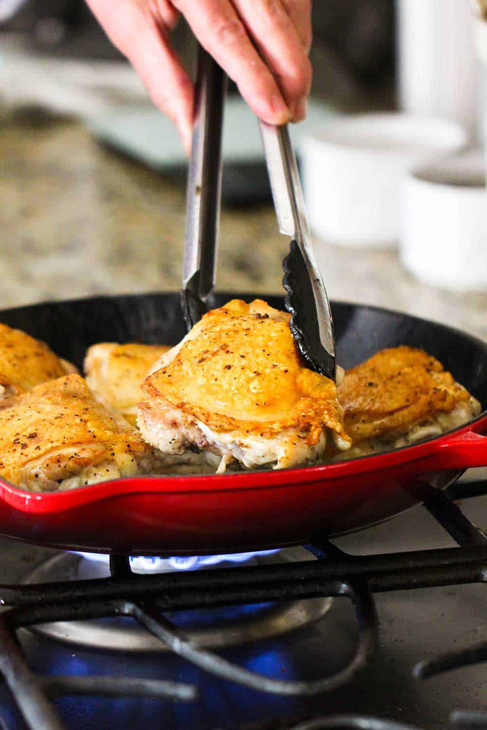 A hand using a pair of tongs to sear chicken thighs in a large red skillet on the stove. 