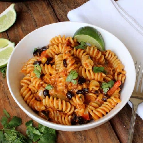 A white bowl holding Easy Tex-Mex Pasta Dinner next to cut limes, fresh cilantro , and a white napking,