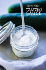 A small jar filled with homemade tzatziki sauce with a small spoon in it, next to two cucumbers and a grey napkin.