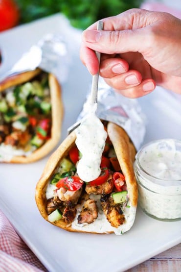 A hand using a small spoon to drizzle homemade tzatziki sauce over the end of a chicken gyro that is sitting next to a small jar of the sauce on a white platter.