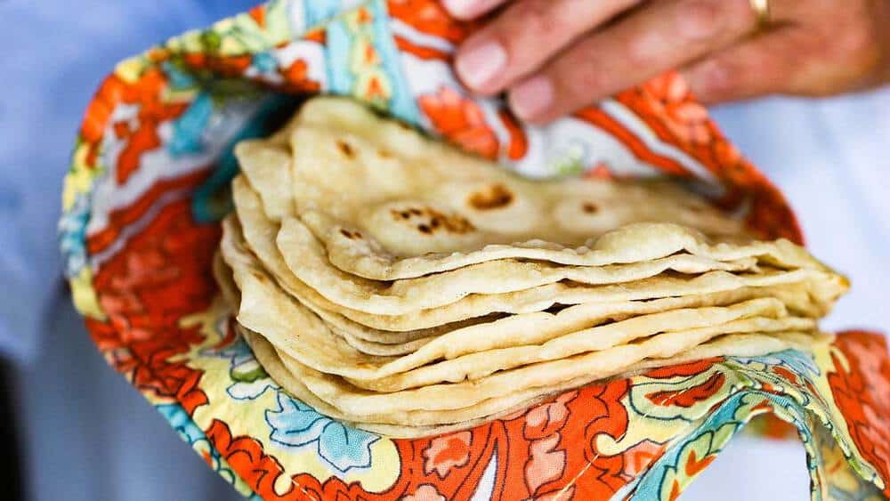 A hand holding a stack of homemade flour tortillas in a colorful cloth. 