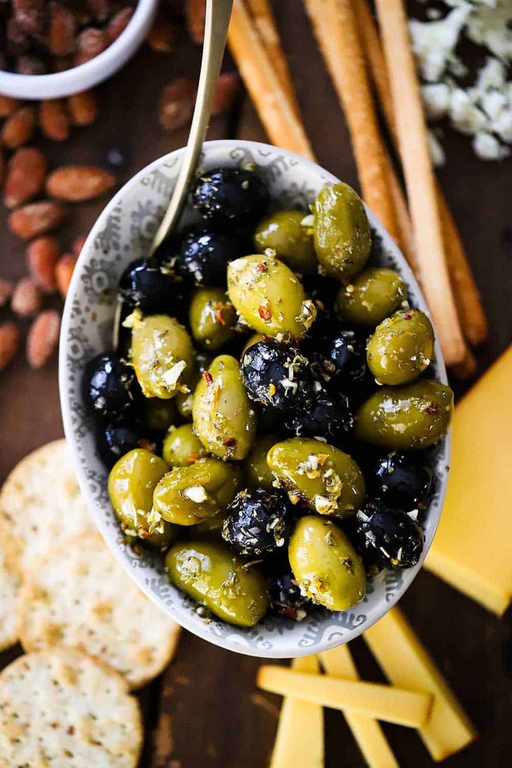 A small oval bowl filled with marinated olives surrounded by cheese slices, crackers, bread sticks, and almonds. 