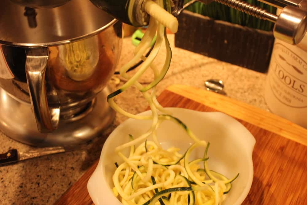 A stand mixer with a spiralizer attachment with spiralized squash and zuchini in a small white bowl. 