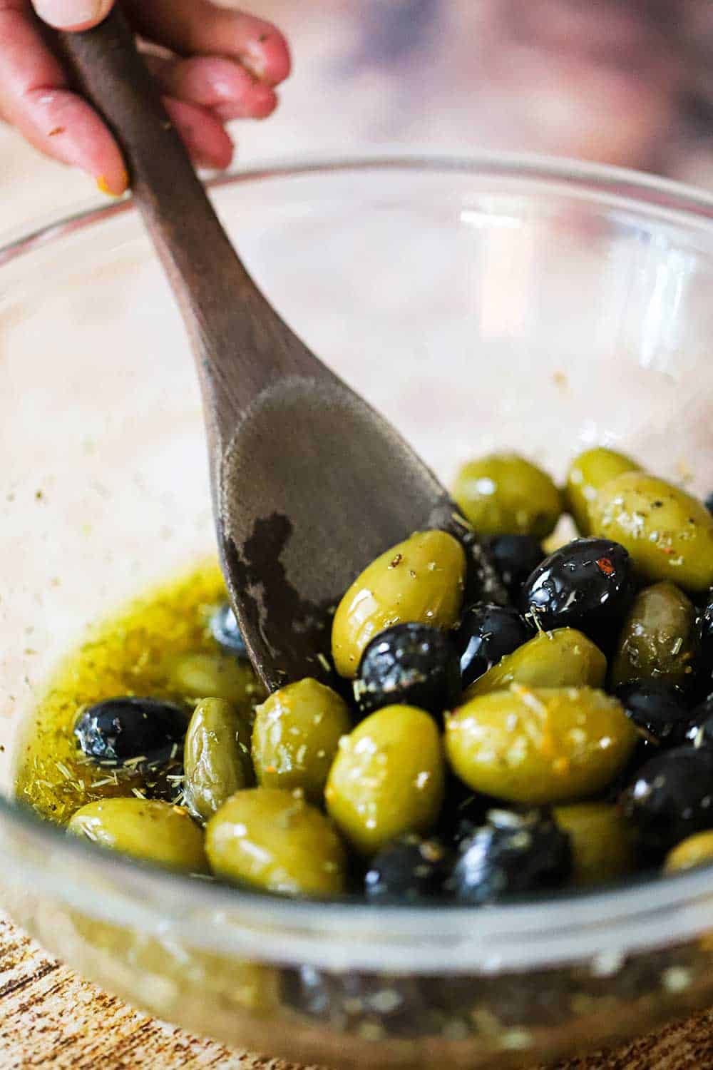 A large glass bowl filled with marinated olives in oil with a wooden spoon stirring them all together. 