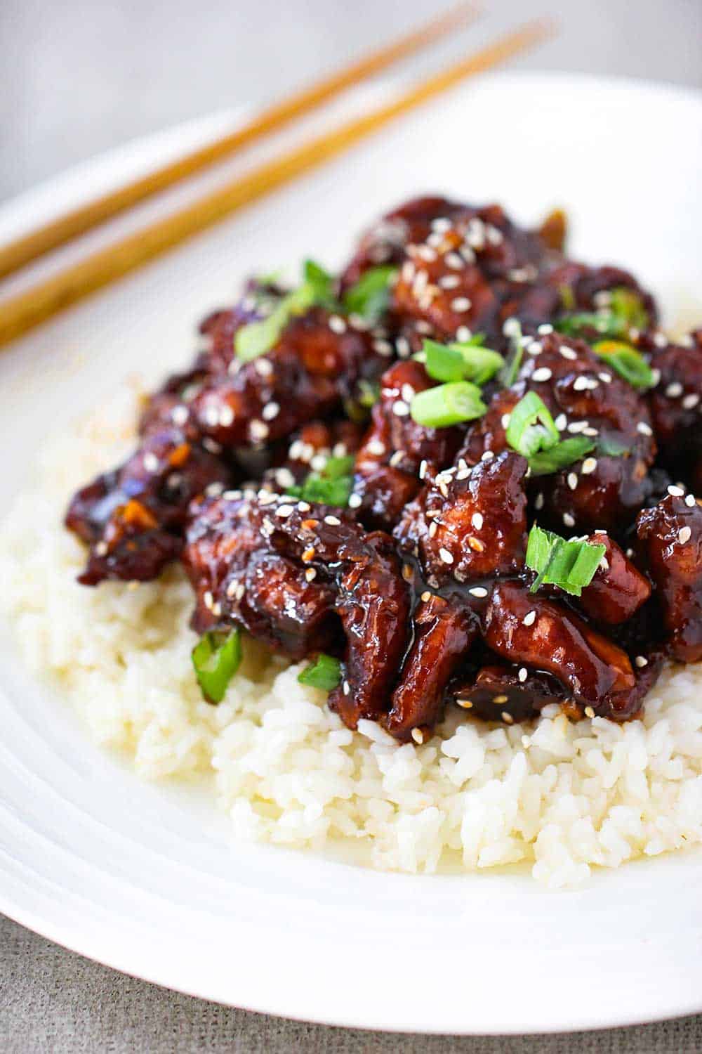 A white plate of General Tso's Chicken on a bed of white rice with a pair of chop sticks nearby. 